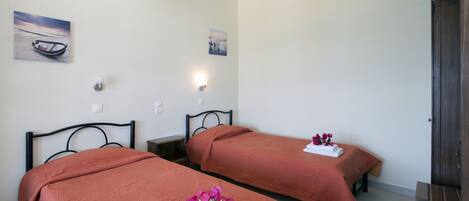 Dos camas individuales con ropa de cama roja a juego, una mesita de noche pequeña, una lámpara de pared y un cuadro de un barco en la pared.