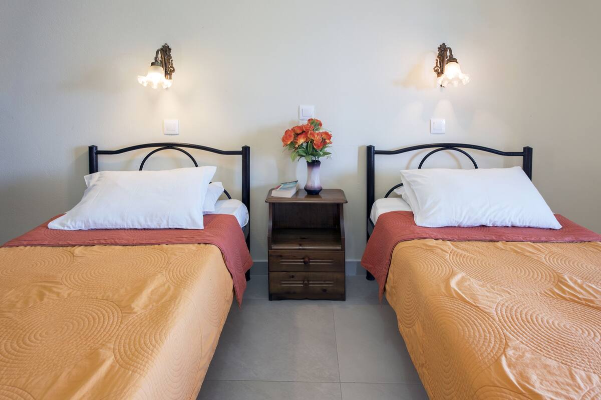 Deux lits simples avec des têtes de lit, une table de chevet ornée d’un vase de fleurs et des lampes murales placées entre les lits.