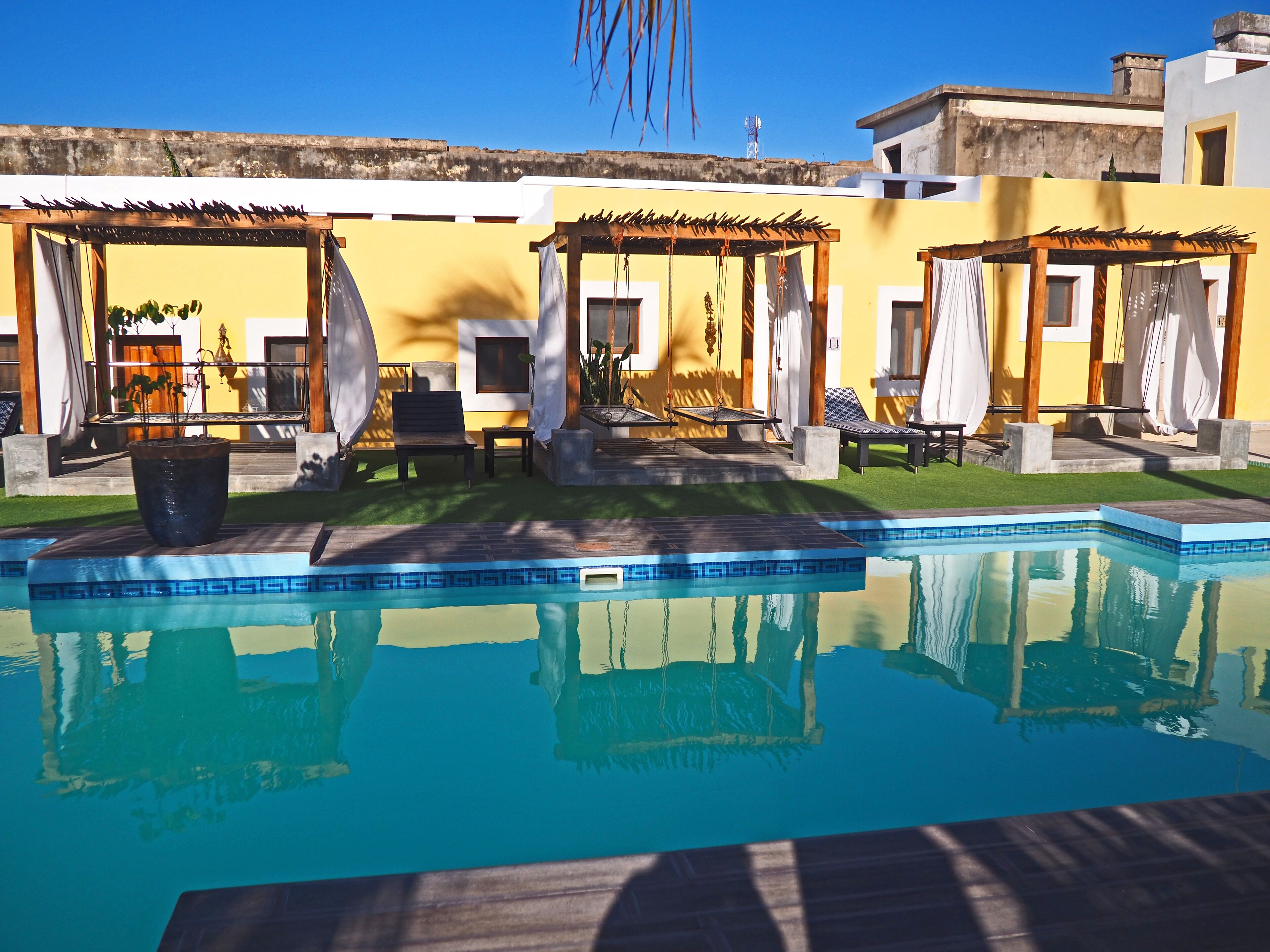 A swimming pool with a pergola and lounge chairs, surrounded by a well-maintained lawn and a yellow building.