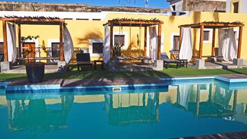 A swimming pool with a pergola and lounge chairs, surrounded by a well-maintained lawn and a yellow building.