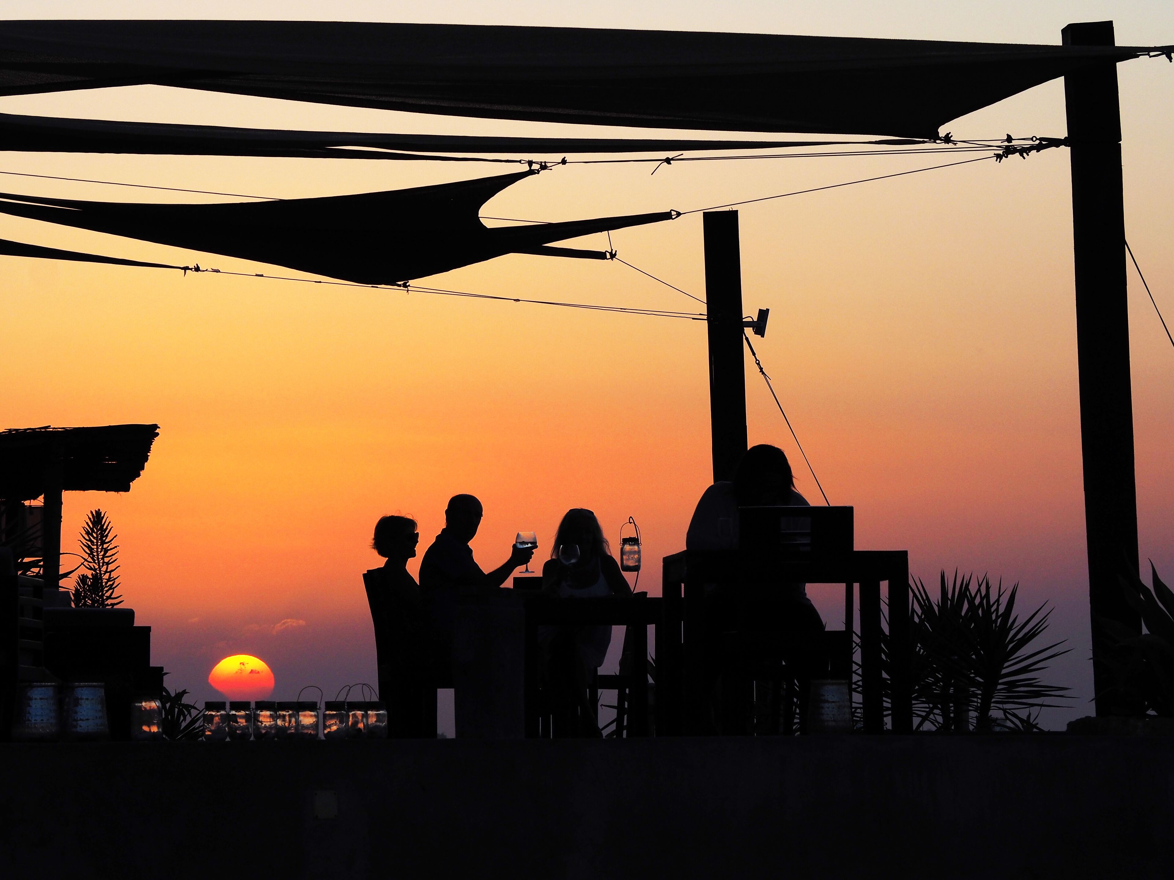 Escena de atardecer con personas siluetas contra el cielo, una mesa con bebidas y un edificio de techo inclinado.