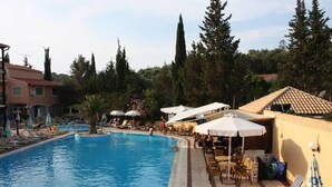 A swimming pool with lounge chairs and umbrellas, surrounded by trees and buildings. - Philippos Apartments (Corfu)