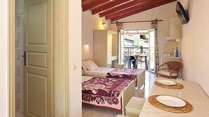 A room with two beds, a dining table with placemats, a TV mounted on the wall, and a balcony with a railing. - Philippos Apartments (Corfu)