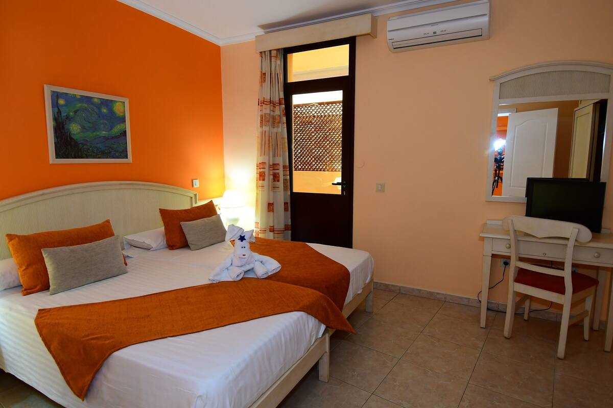 Habitación de hotel con dos camas, un escritorio, una silla, un cuadro y una puerta con vistas.