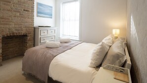 A bedroom with a brick fireplace, a bed with white bedding, a nightstand with a lamp, and a window with blinds. - Sail Away - 2-Bed Apartment by the Beach (Whitstable)
