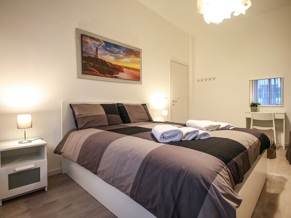 A bedroom with a bed, bedside tables, a painting, a desk, and a chair.