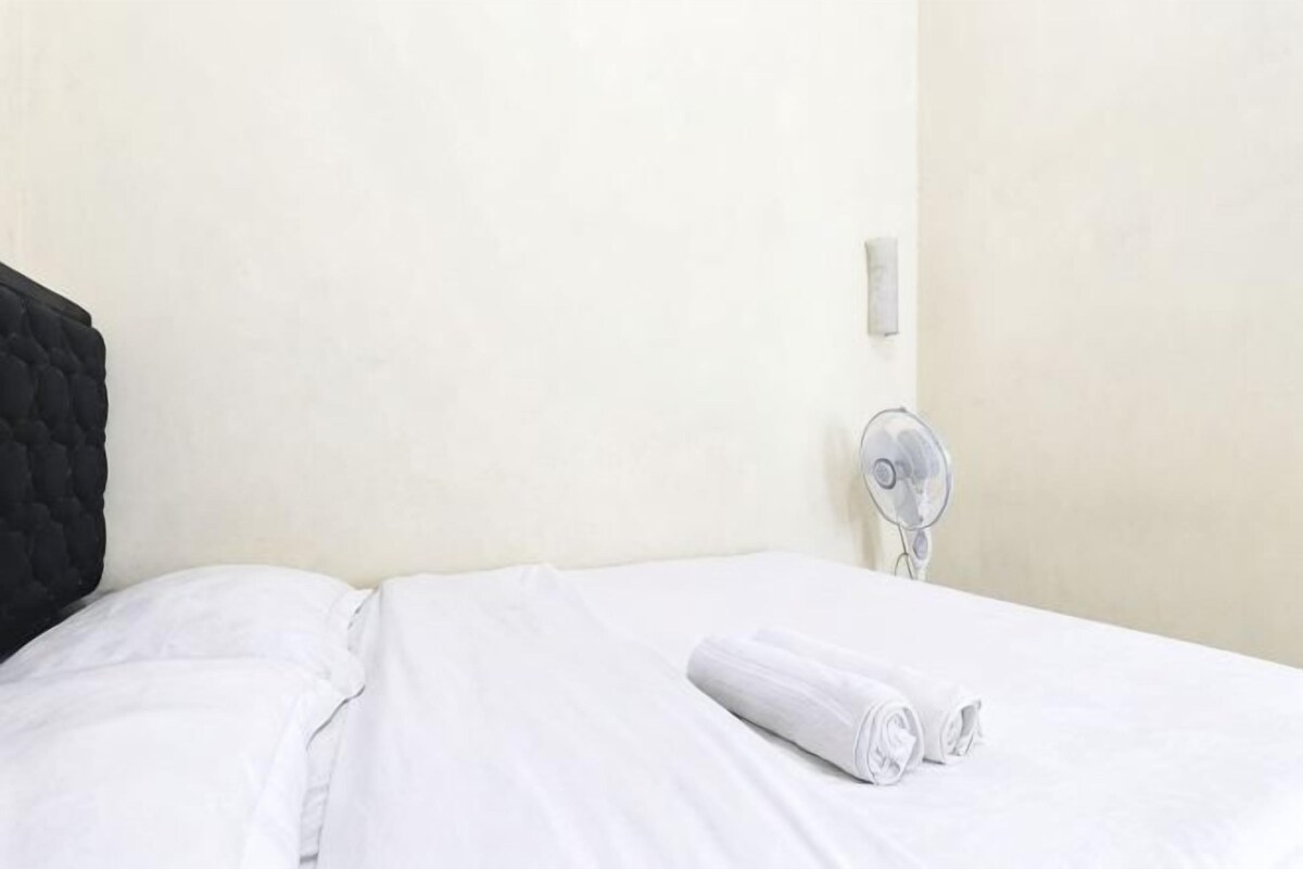 Een bed met witte lakens en een opgevouwen handdoek, een zwart hoofdeinde en een ventilator bevestigd aan de muur.