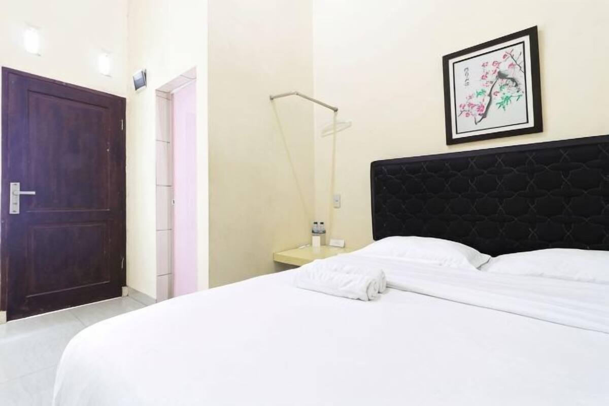 A hotel room with a bed, a door, a nightstand with a lamp, and a framed picture on the wall.