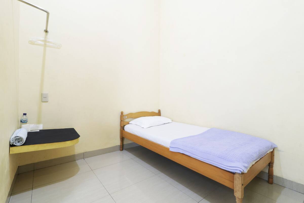 A small, single-bed room with a wooden bed, a small bench with a towel, and a wall-mounted light fixture.