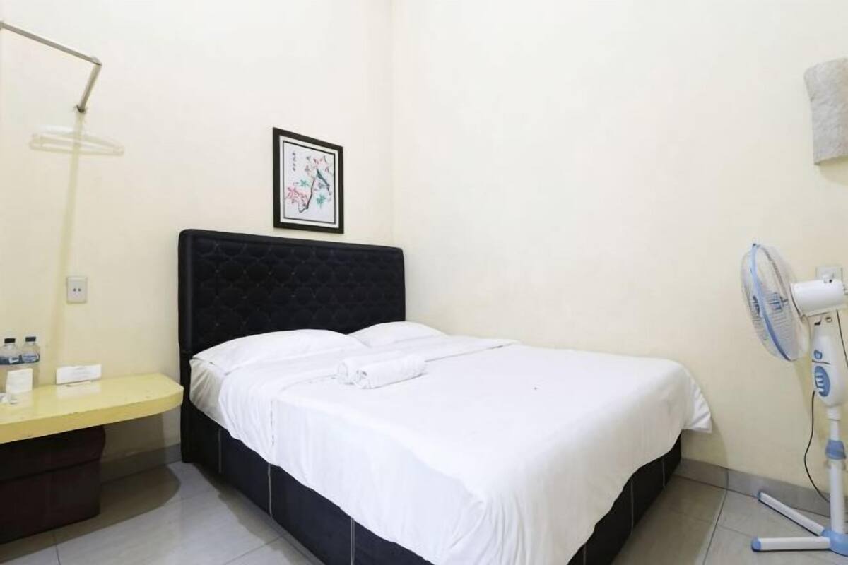 A hotel room with a bed, a bedside table with a lamp, a wall-mounted picture, and a standing fan.