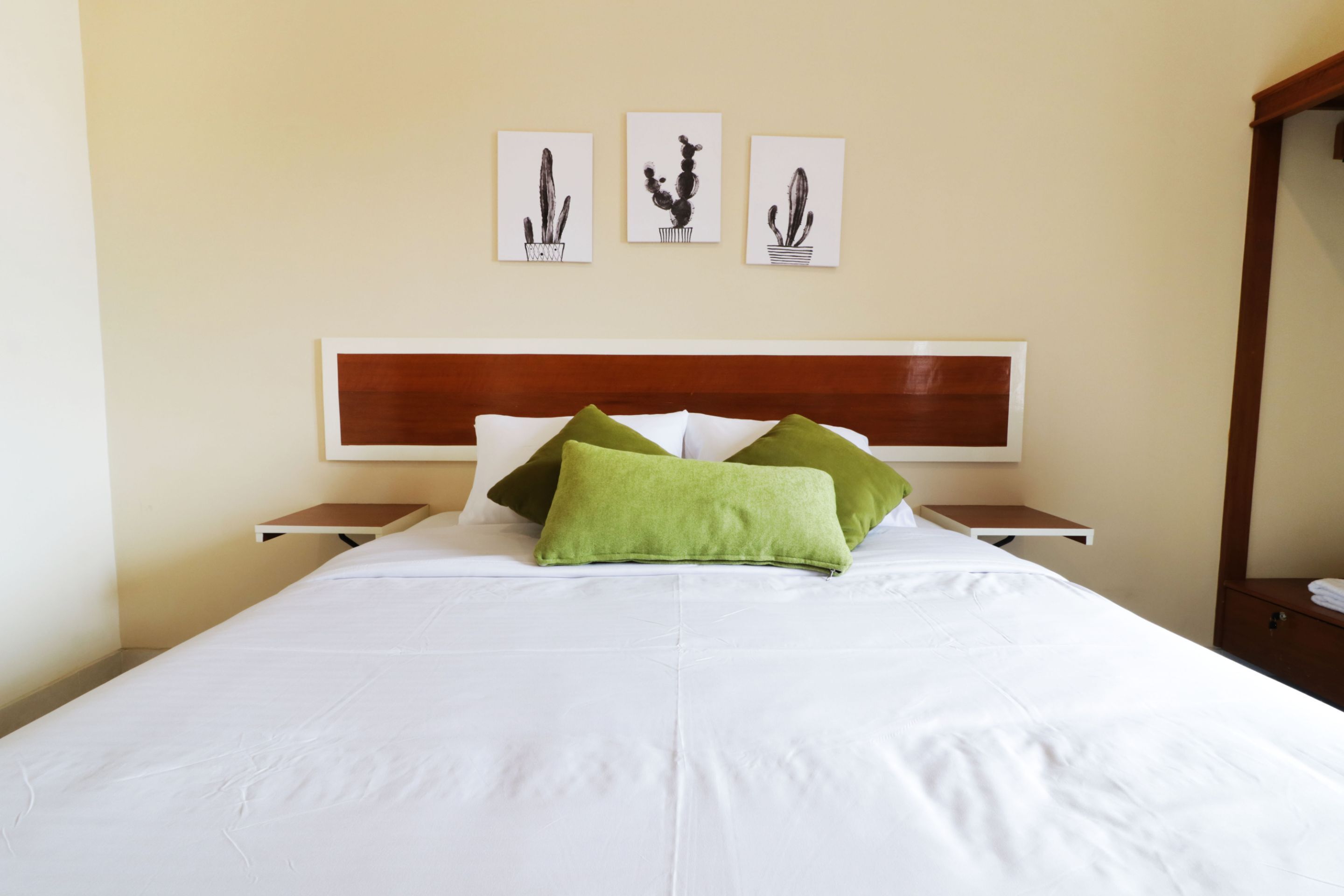 A neatly made bed with white linens and green pillows, a wooden headboard, and three framed cactus artworks on the wall.