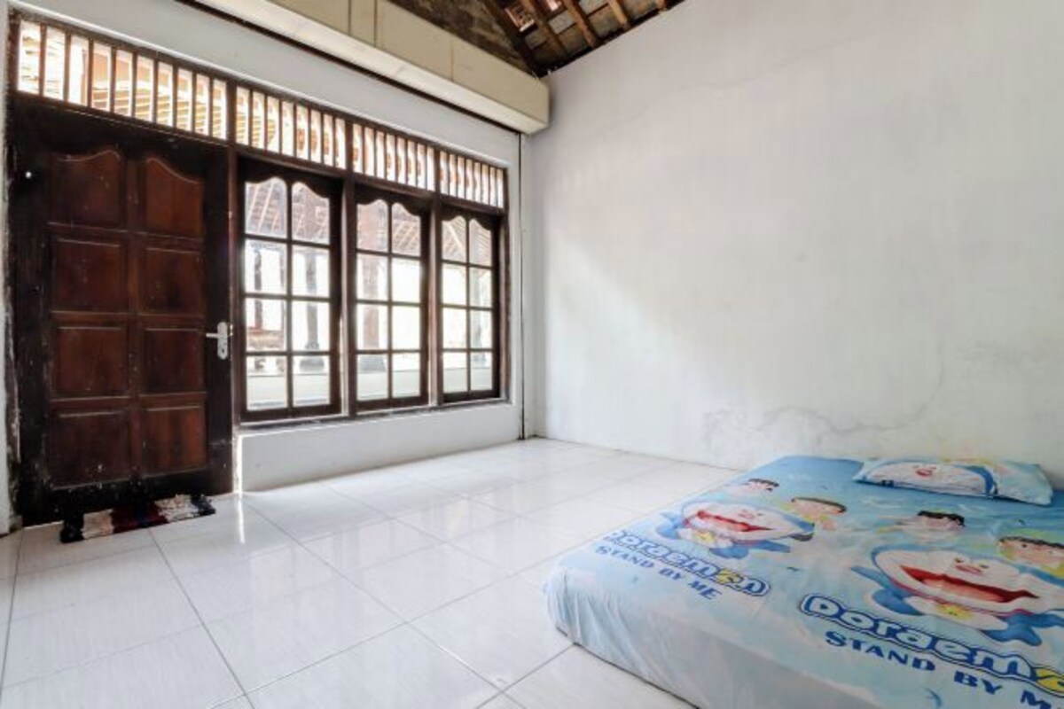 Ein Zimmer mit großem Fenster, einem Bett mit Doraemon-Bettdecke und einer Holztür.
