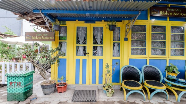 Un edificio azul y amarillo con un letrero que dice 'Home Stay'. Hay plantas en macetas, un bote de basura verde y dos sillas de mimbre afuera.