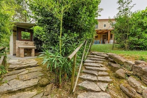 A stone pathway leading to a brick structure with a window, surrounded by lush greenery and a house in the background.