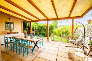 An outdoor patio with a wooden table set for dining, surrounded by blue chairs, and a hanging chair.