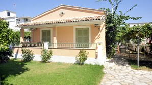 A two-story building with a tiled roof, a balcony with a railing, and a small garden area with grass and shrubs. - Kipos Apartments (Corfu)