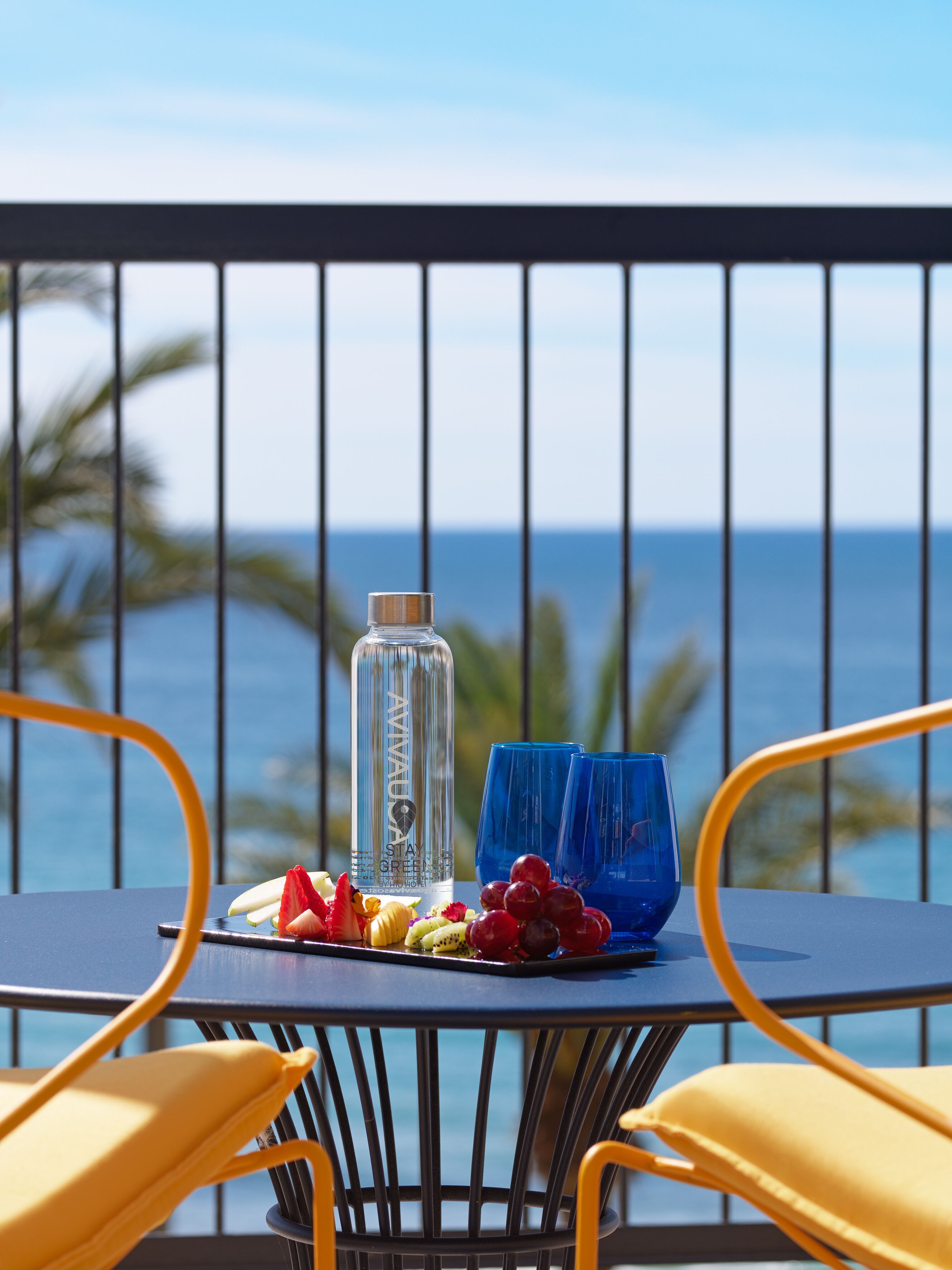 A table with a bottle, blue glasses, and fruit on a balcony with a view of the ocean.