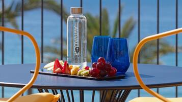 A table with a bottle, blue glasses, and fruit on a balcony with a view of the ocean.