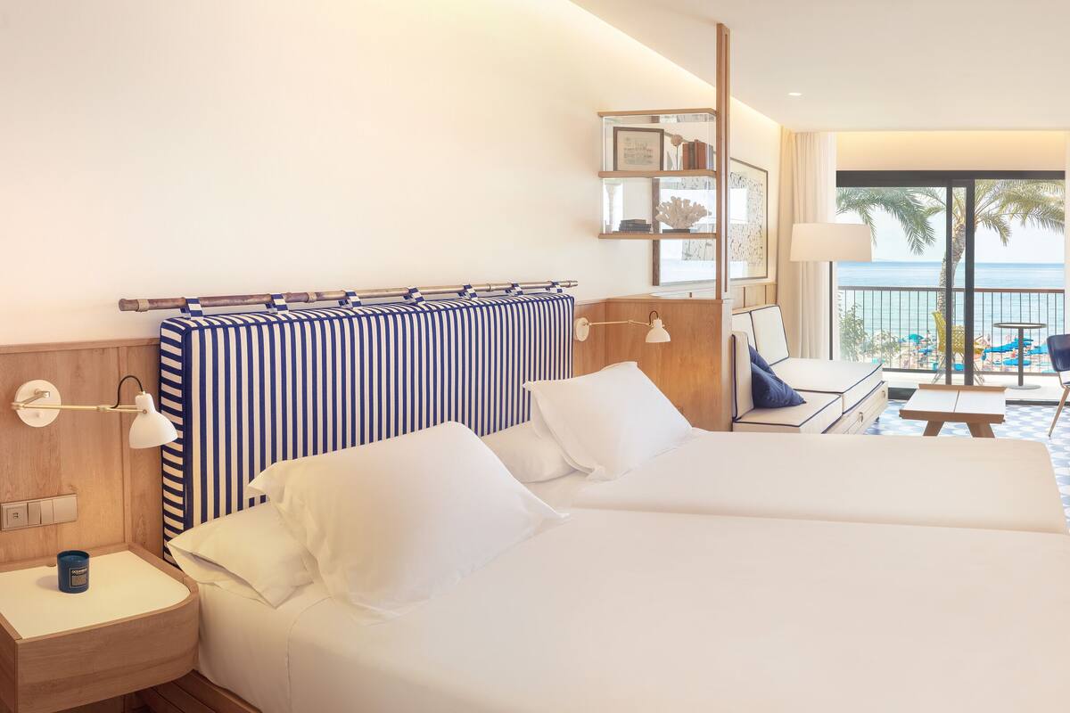 A hotel room with a large bed, a desk with a chair, a small table, a lamp, and a view of the beach and palm trees through a balcony.