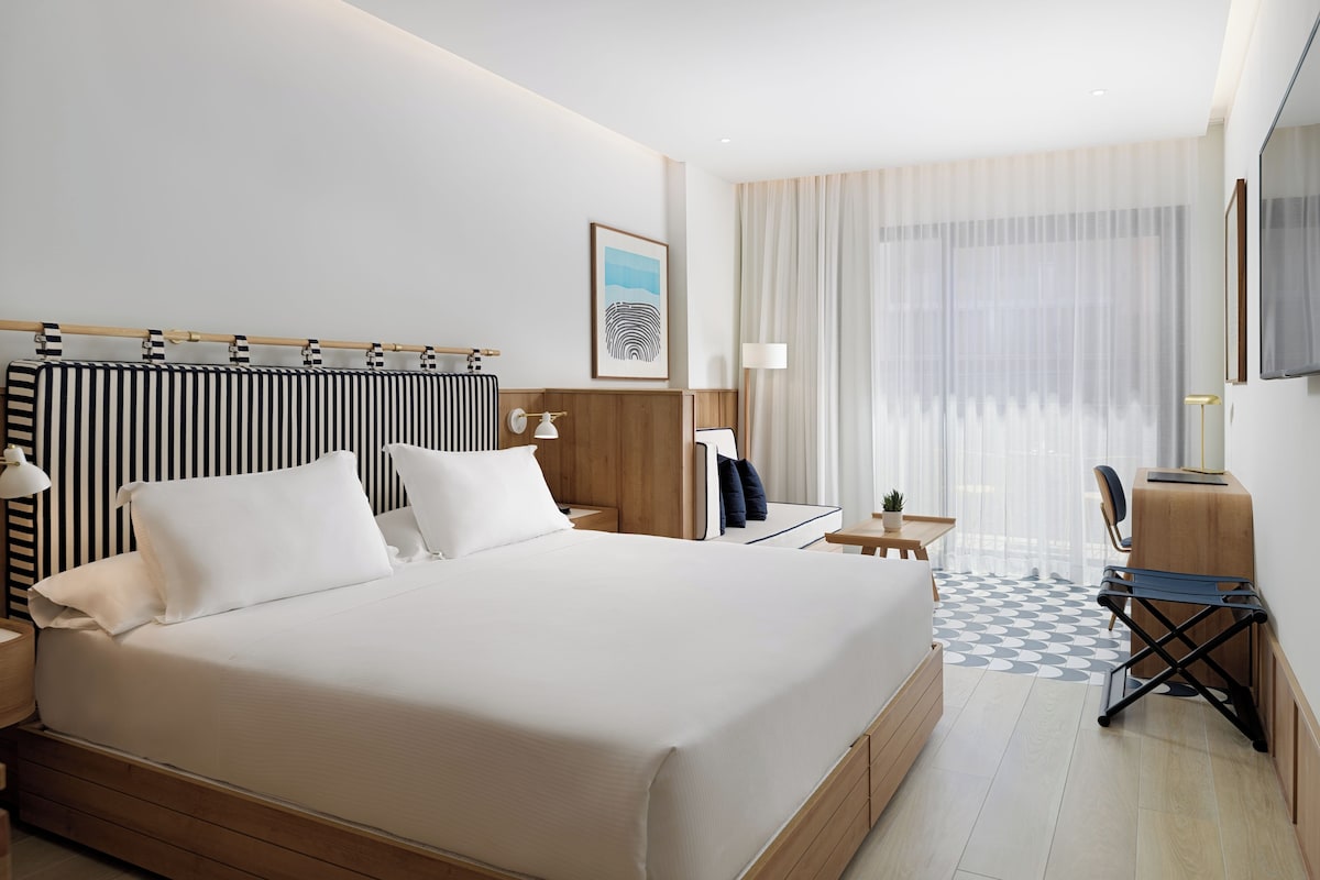 Une chambre d’hôtel avec un grand lit, un bureau, une chaise, une lampe et une fenêtre avec des rideaux légers.