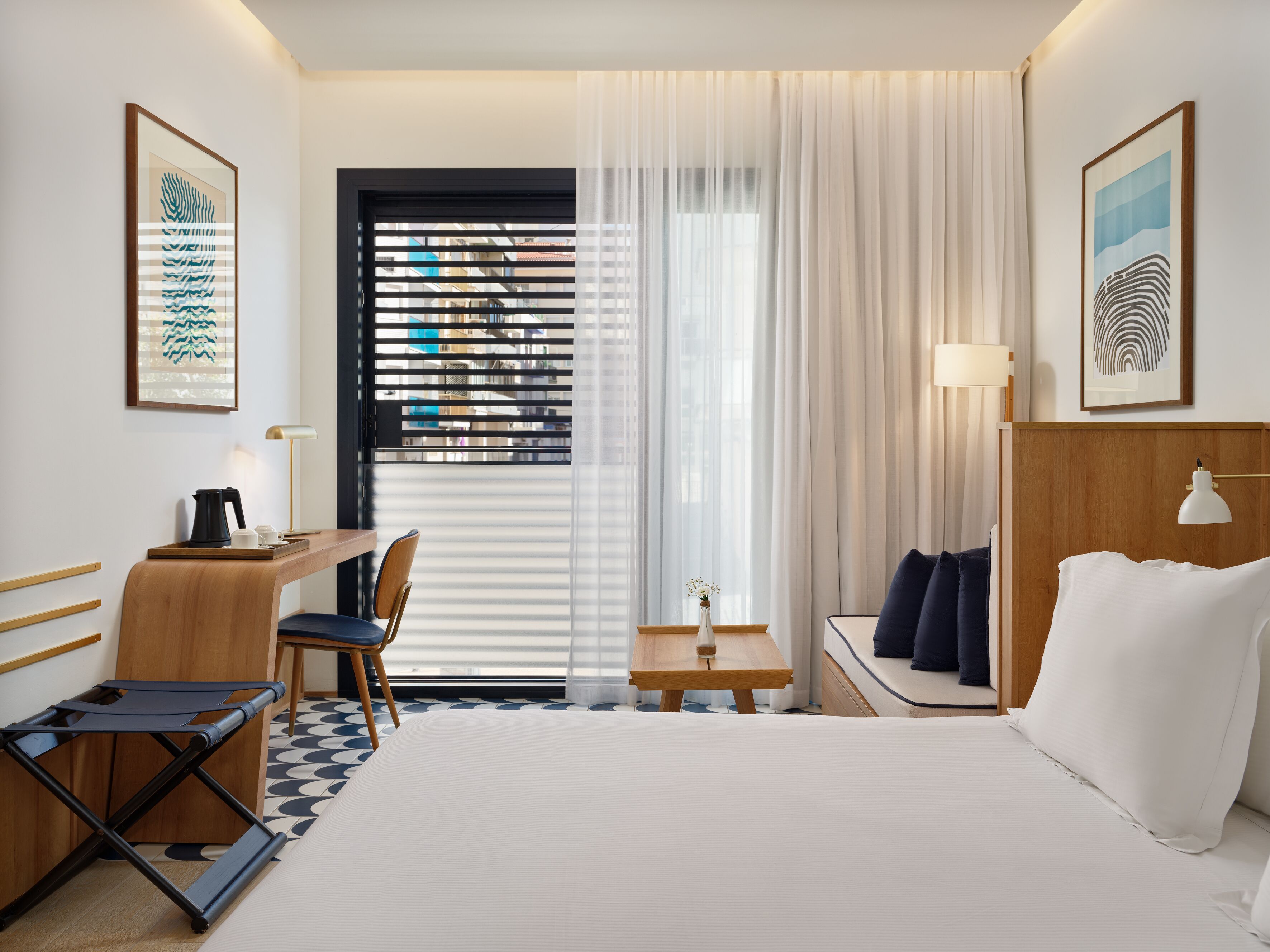 A hotel room with a bed, a desk, a chair, a small table with a vase, and a window with blinds.