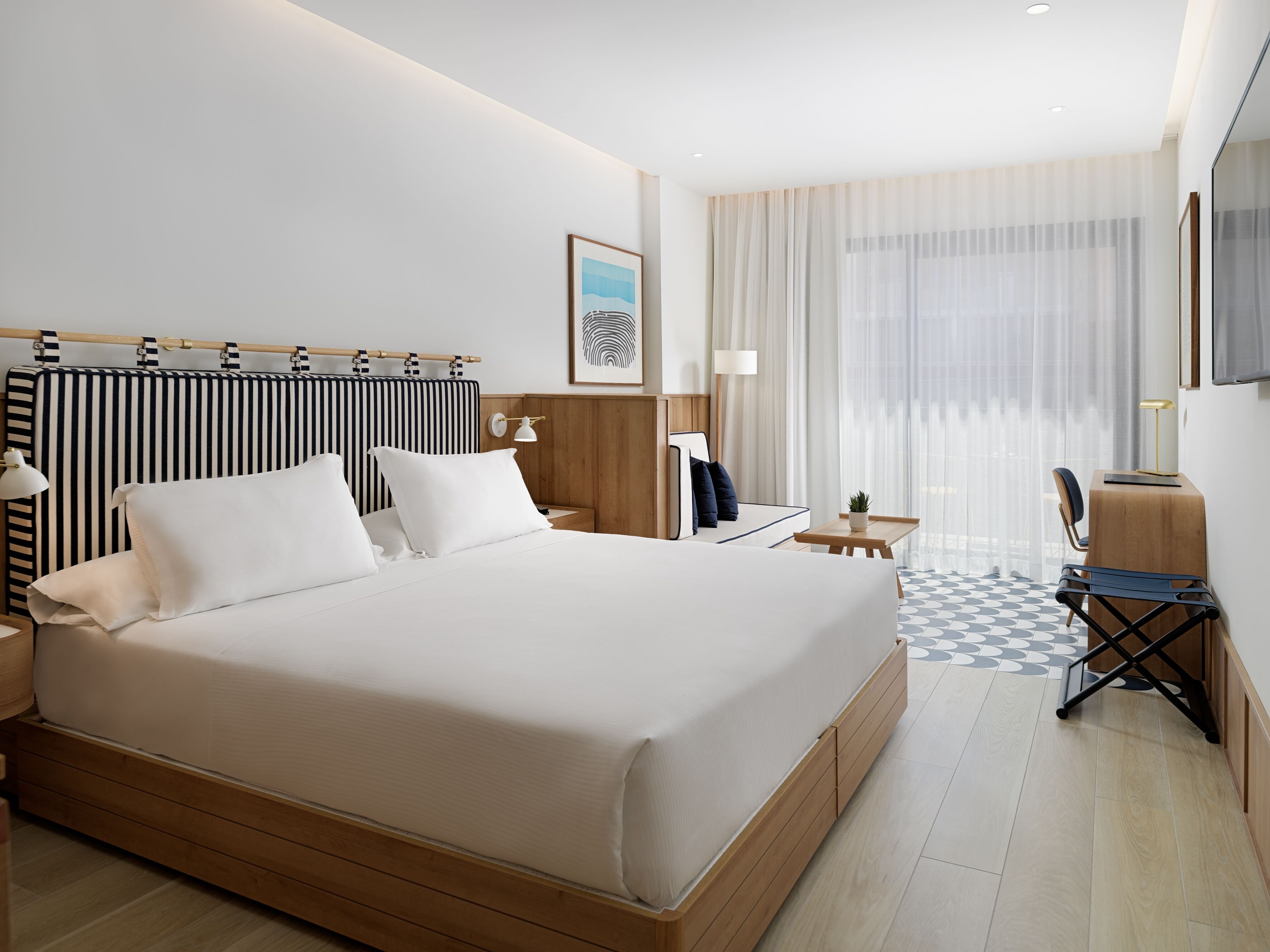 Una habitación de hotel con una cama grande, un escritorio, una silla, una lámpara y una ventana con cortinas translúcidas.
