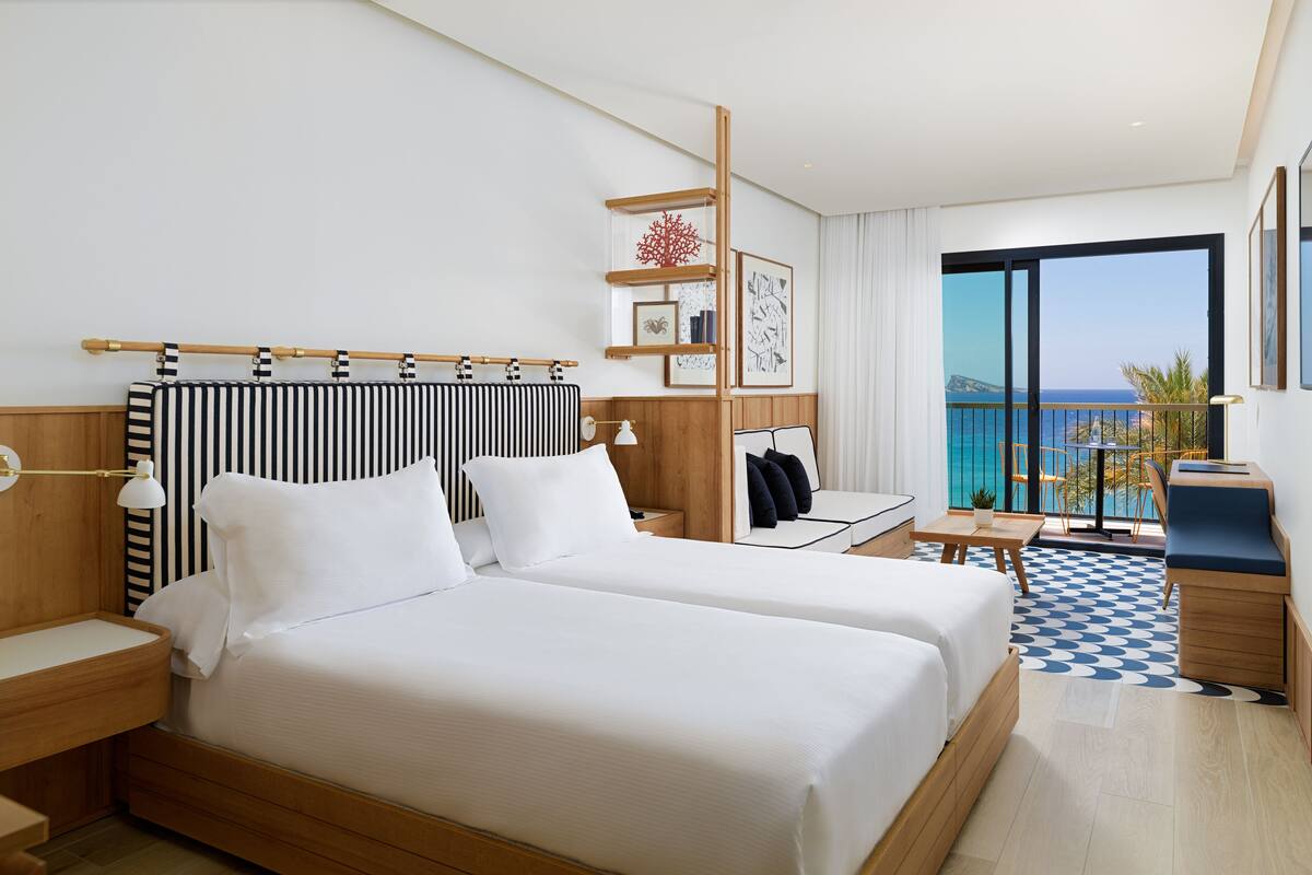 A hotel room with a large bed, a small table, a chair, a desk, and a balcony with a view of the sea.