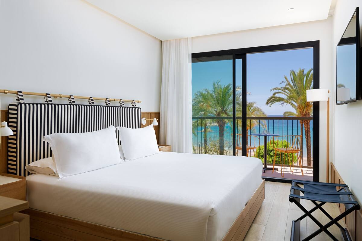 A hotel room with a large bed, a wooden headboard, a balcony with a view of the sea, and a TV mounted on the wall.