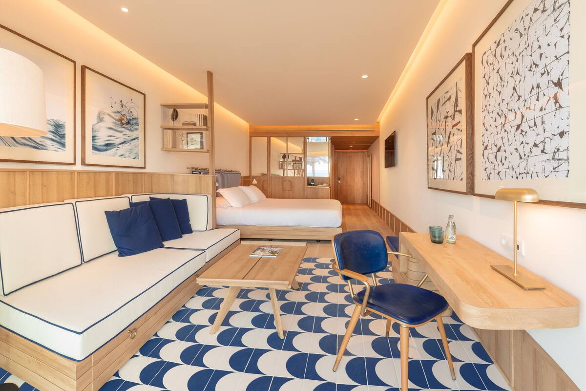 A modern hotel room with a sofa, a bed, a desk, and a chair. There are framed pictures on the walls and a patterned floor.