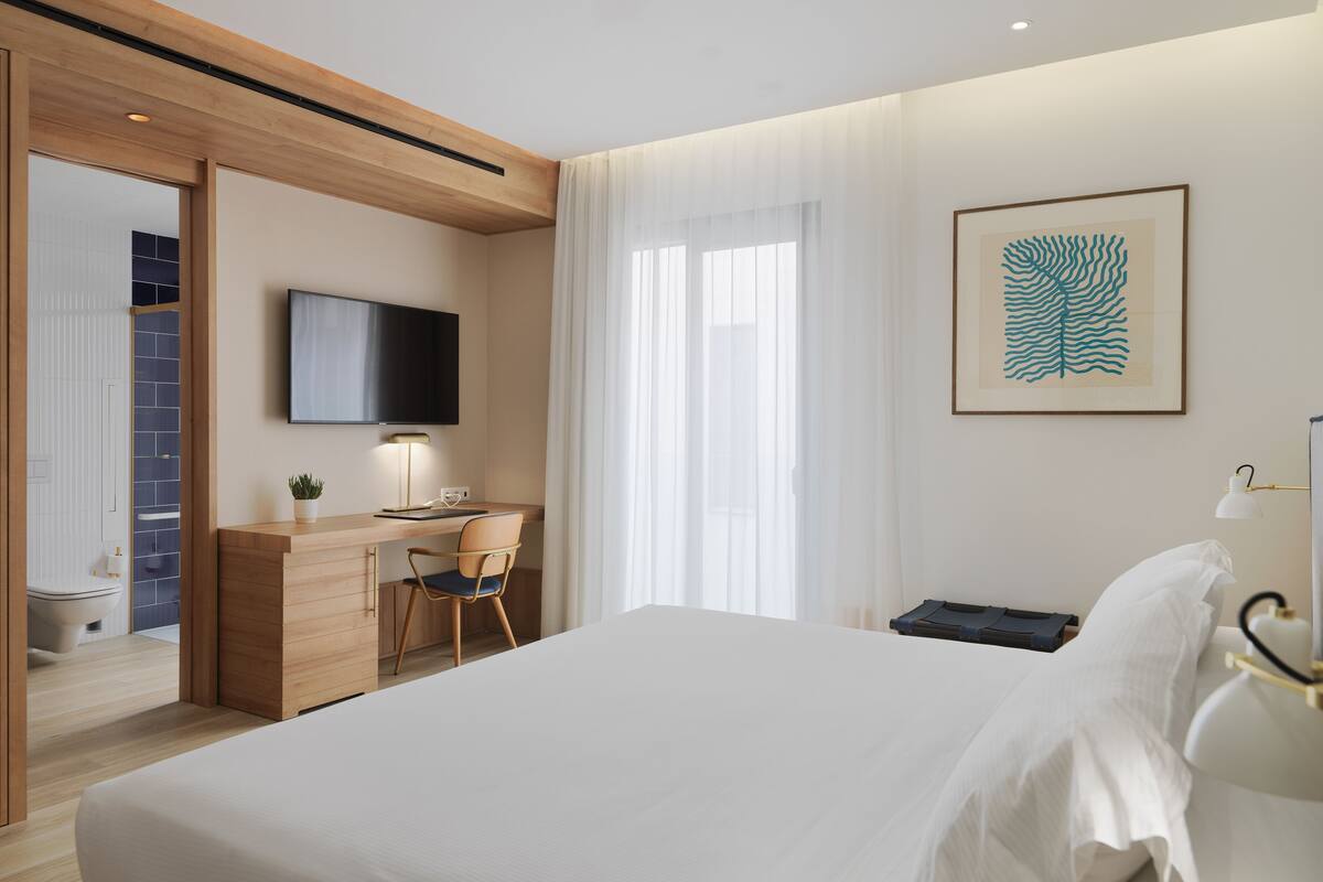 A modern hotel room with a large bed, a desk with a chair, a television, and a bathroom visible through an open door.