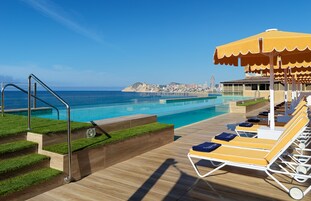 A poolside area with sun loungers, a wooden deck, and a view of the sea and cityscape.