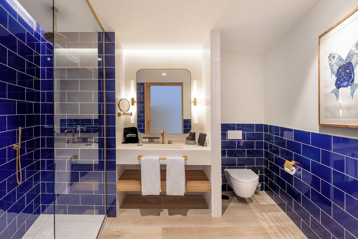 Une salle de bain moderne avec des carreaux bleus, une douche en verre, un lavabo blanc avec un robinet doré et une œuvre d’art encadrée au mur.