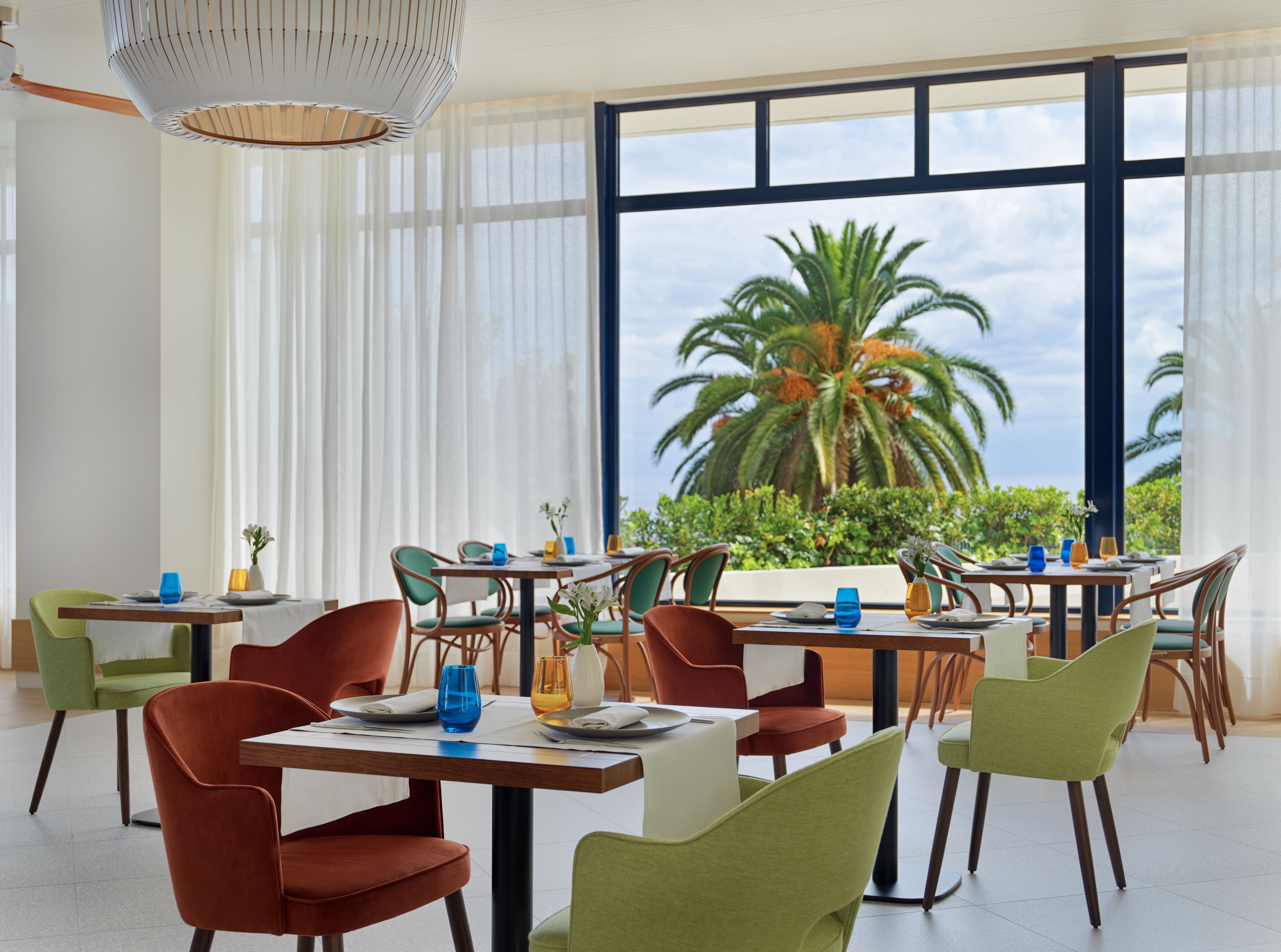 Un espace repas avec des tables et des chaises, de grandes fenêtres offrant une vue sur les palmiers et le ciel.