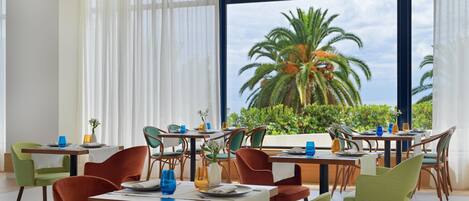 Un área de comedor con mesas y sillas, ventanas amplias con vista a palmeras y al cielo.