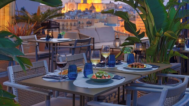 Un área de comedor al aire libre con mesas preparadas para dos personas, con vistas a un paisaje urbano de fondo.