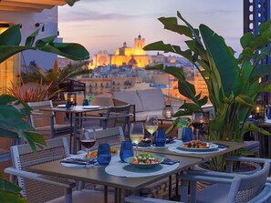 Un área de comedor al aire libre con mesas preparadas para dos personas, con vistas a un paisaje urbano de fondo.