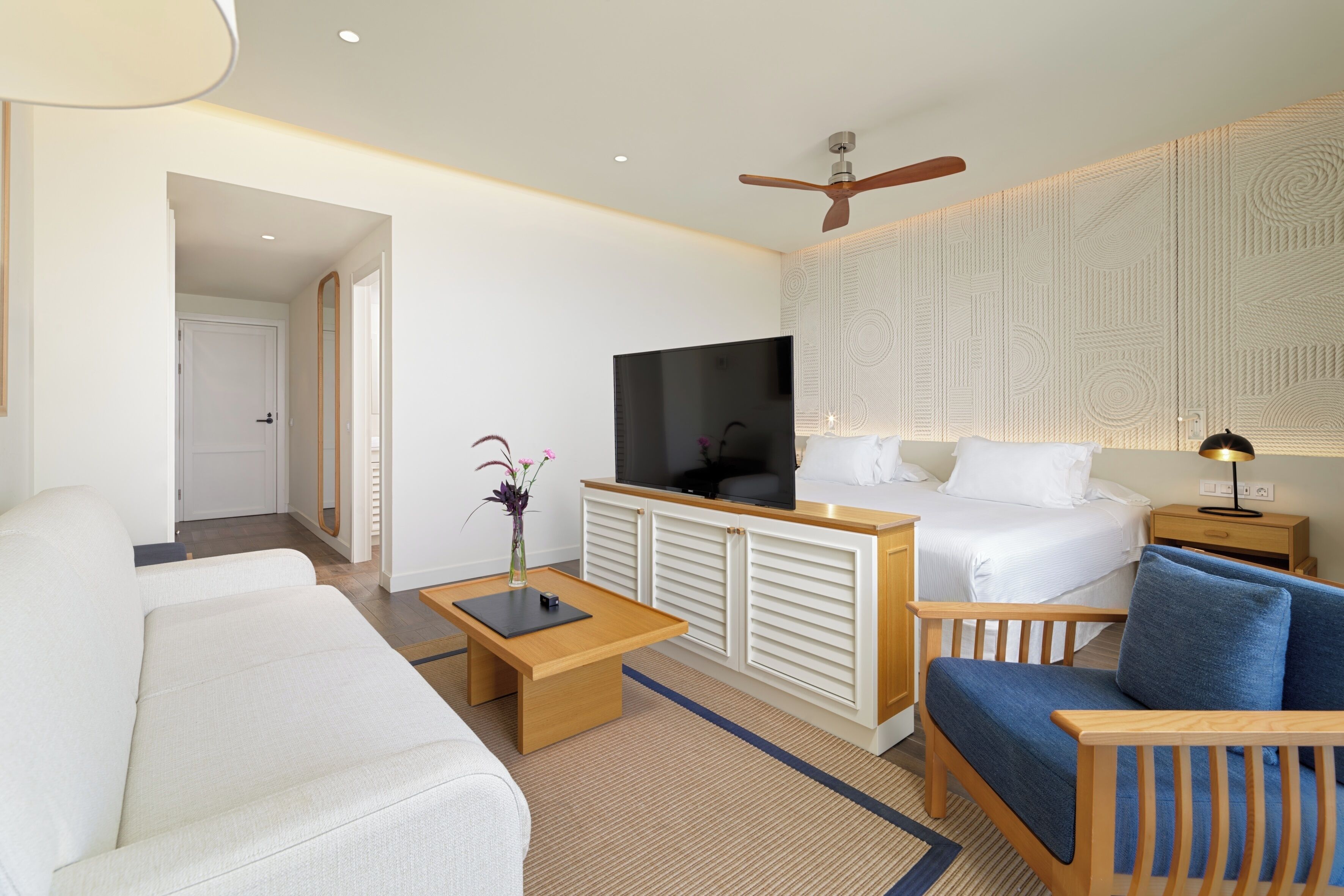 Habitación de hotel con cama, sofá, sillón, televisor y ventilador de techo.
