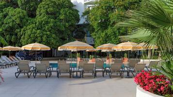 A poolside area with lounge chairs and striped umbrellas, surrounded by lush greenery and a building in the background.