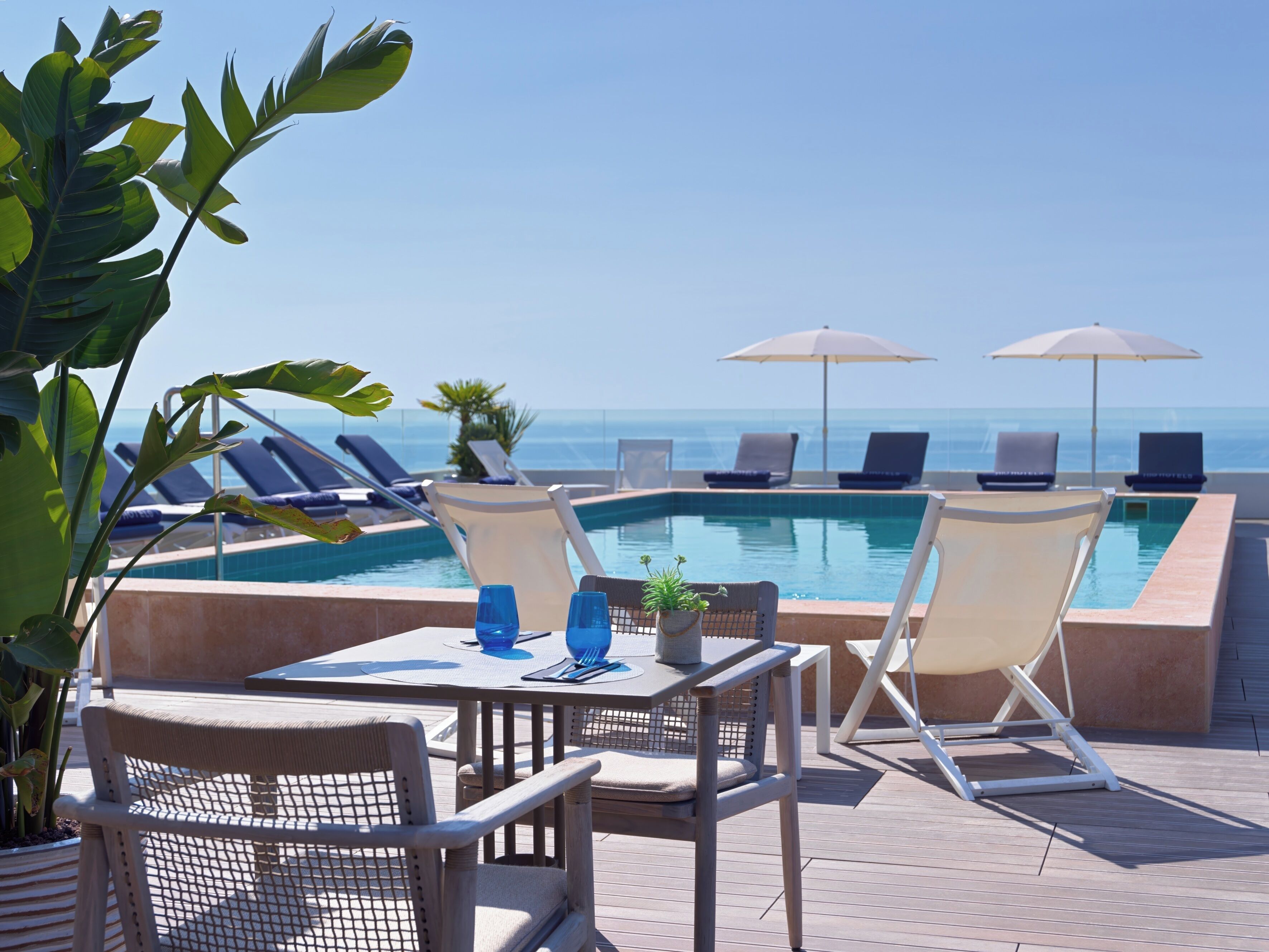 Un espace aménagé au bord de la piscine, avec des chaises longues, une table où sont servis des boissons et des parasols offrant une vue sur l’océan.