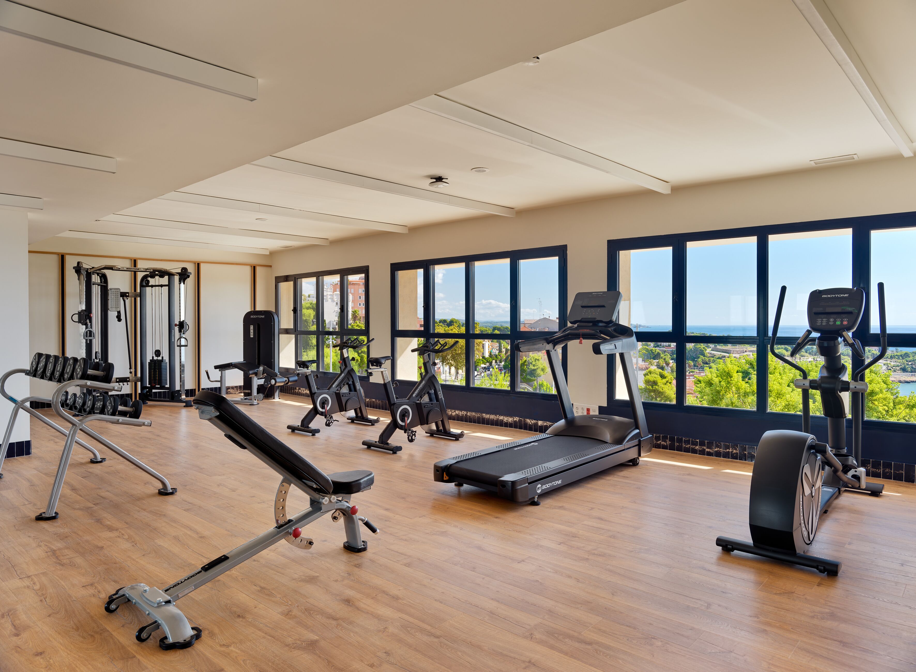 Un gimnasio con máquinas de cardio, equipo de levantamiento de pesas y vistas a zonas verdes y edificios a través de amplias ventanas.