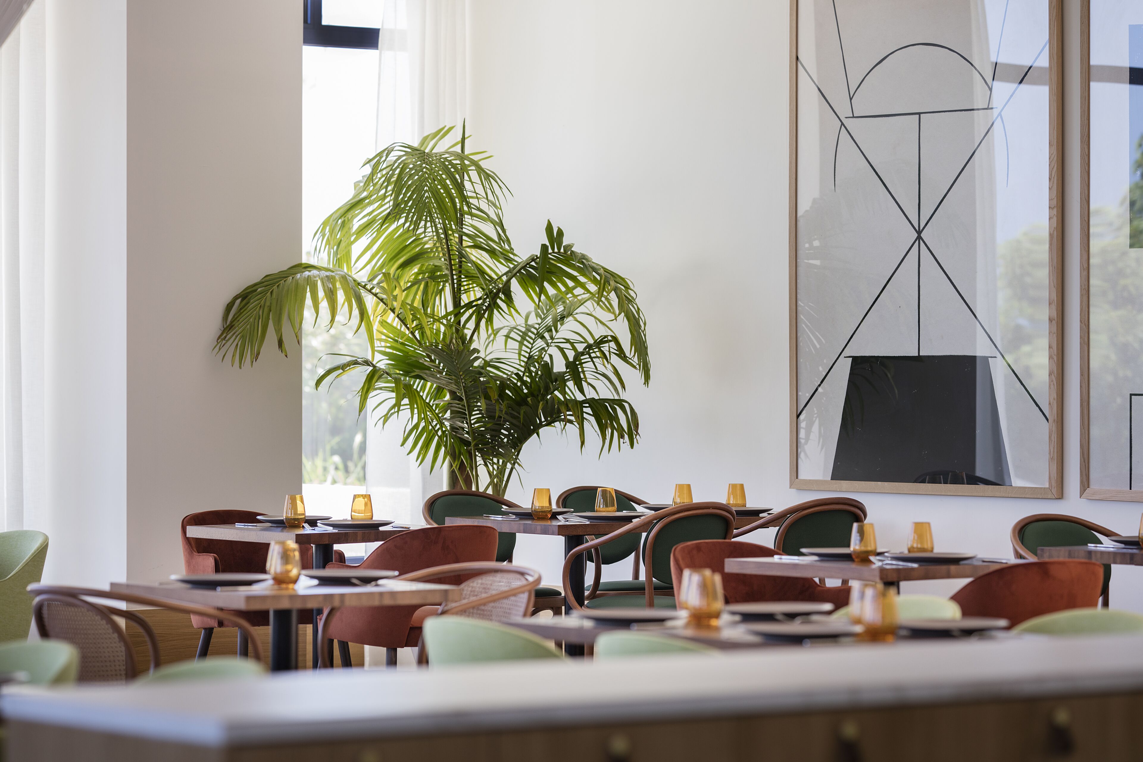 Een restaurant met meerdere tafels en stoelen, een grote potplant en een modern abstract schilderij aan de muur.