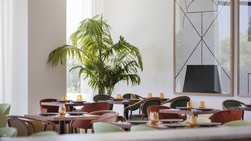 A restaurant with multiple tables and chairs, a large potted plant, and a modern abstract painting on the wall.