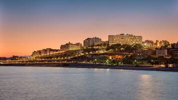 A coastal cityscape at dusk with a prominent building on a hill, a bridge, and a calm sea.