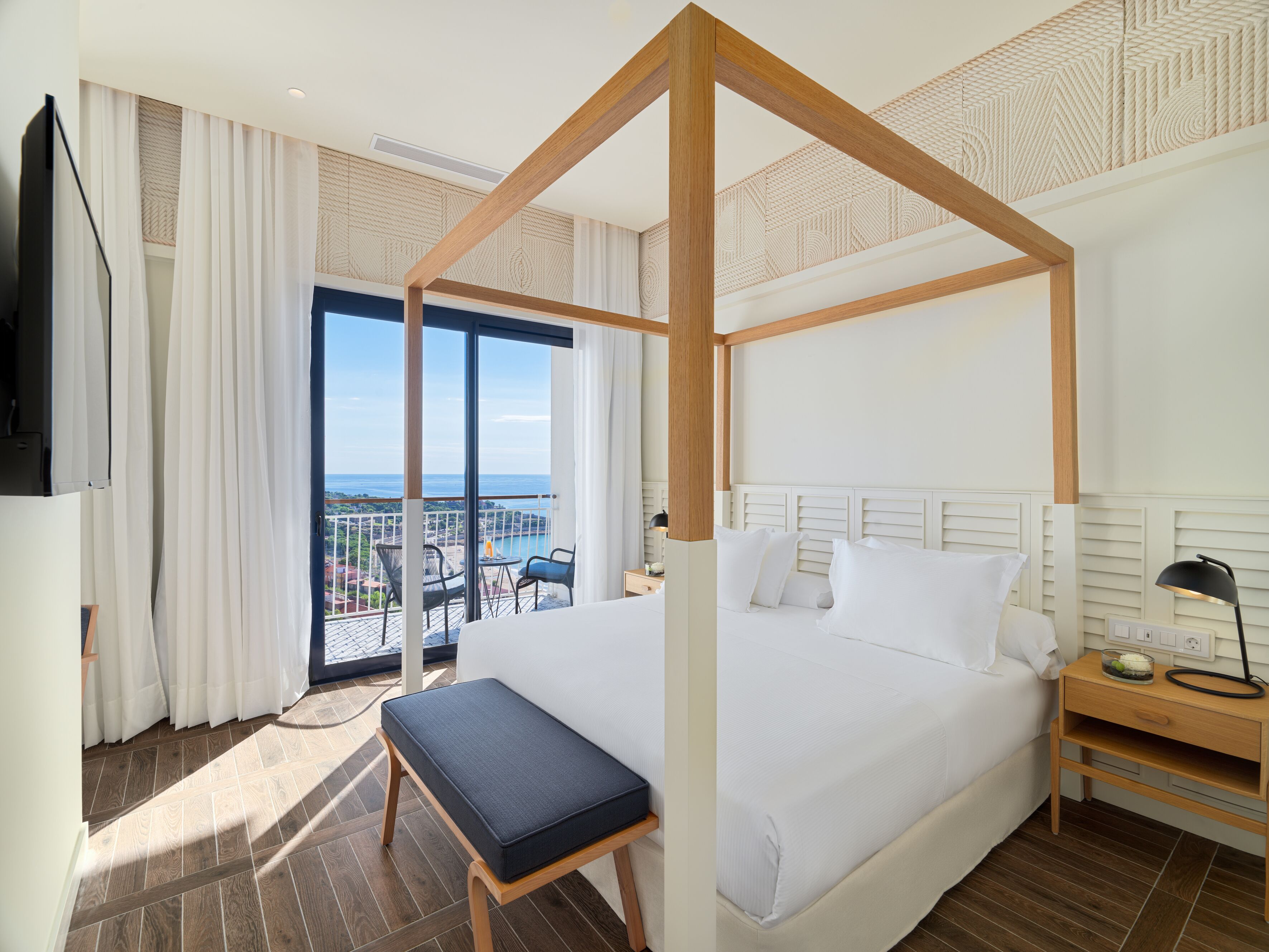 Una habitación de hotel moderna con una cama grande, un dosel de madera, un balcón con vistas y una pequeña mesa con lámpara.