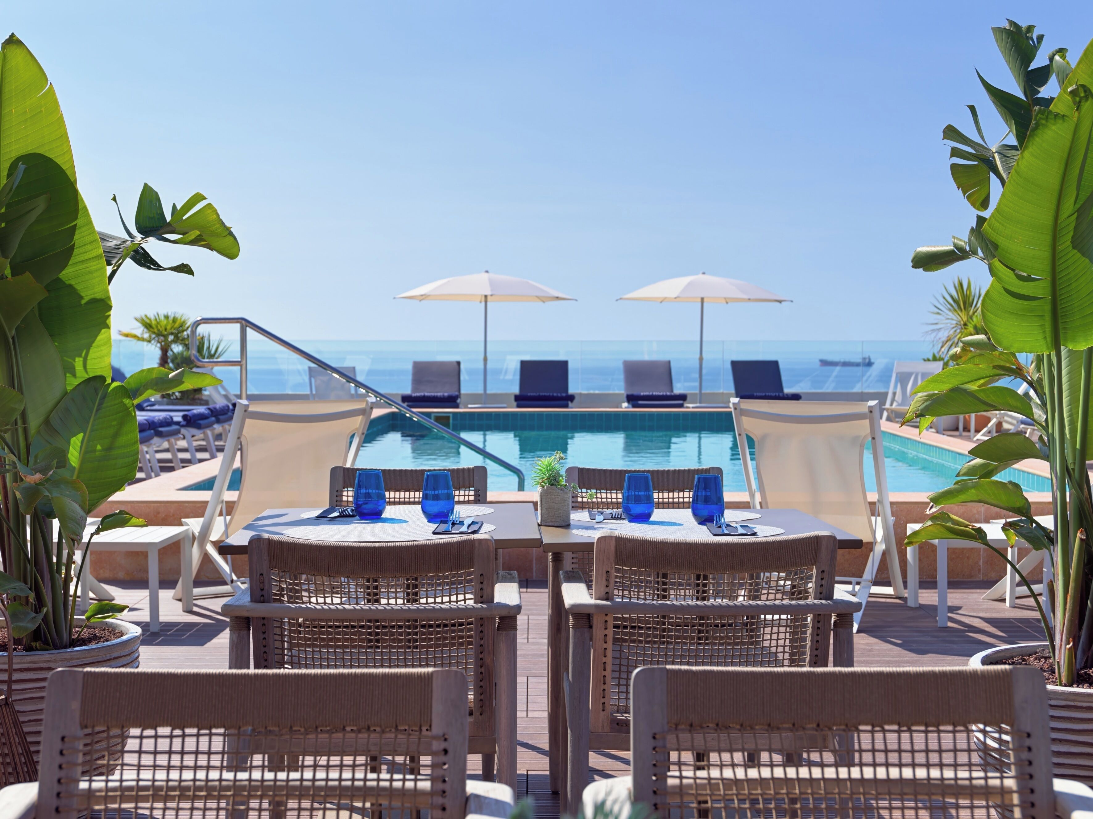 A poolside area with lounge chairs and tables, a pool, and a view of the ocean.