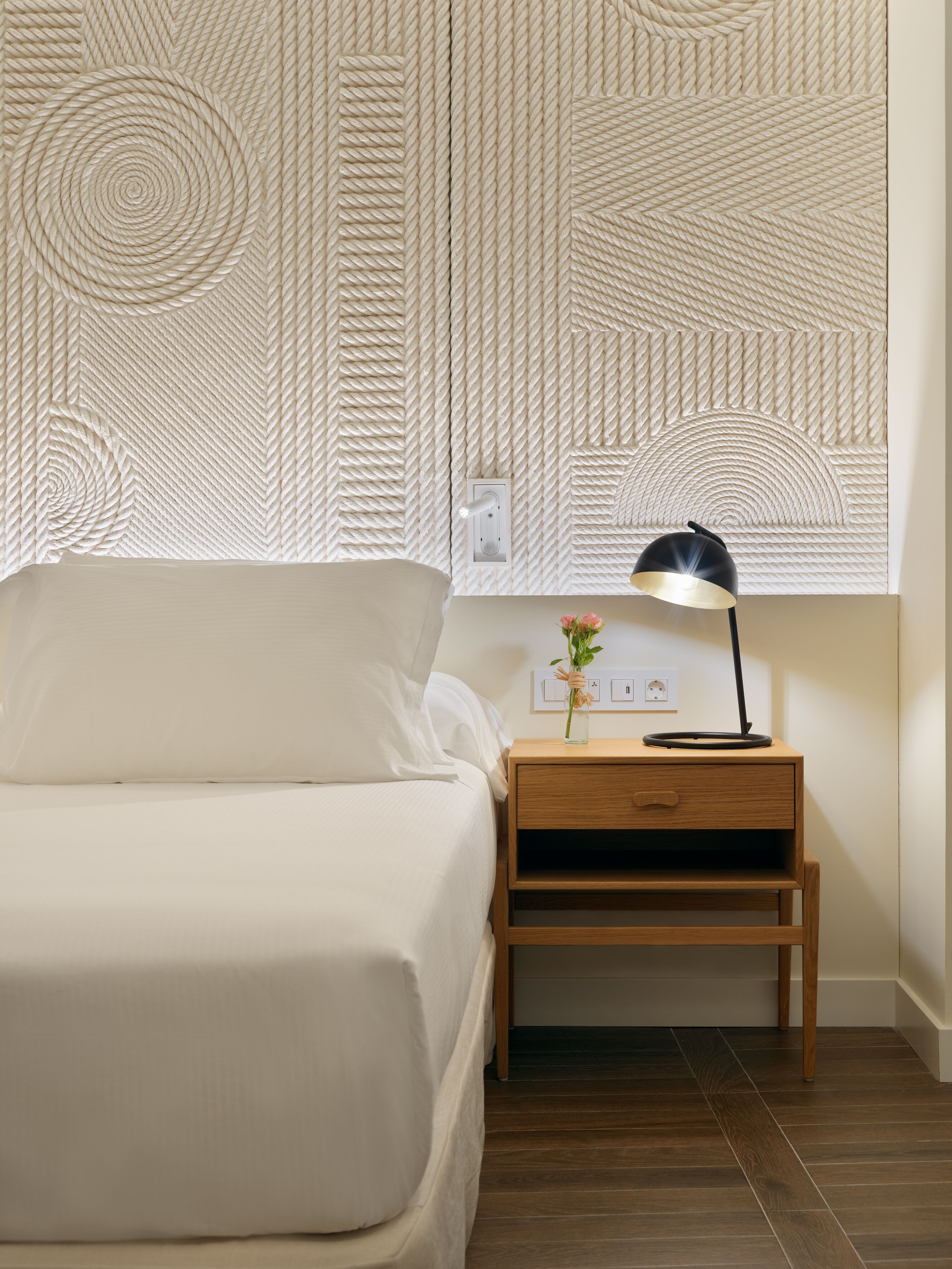 A neatly made bed with a wooden nightstand beside it, a wall-mounted lamp, and a small vase with flowers.