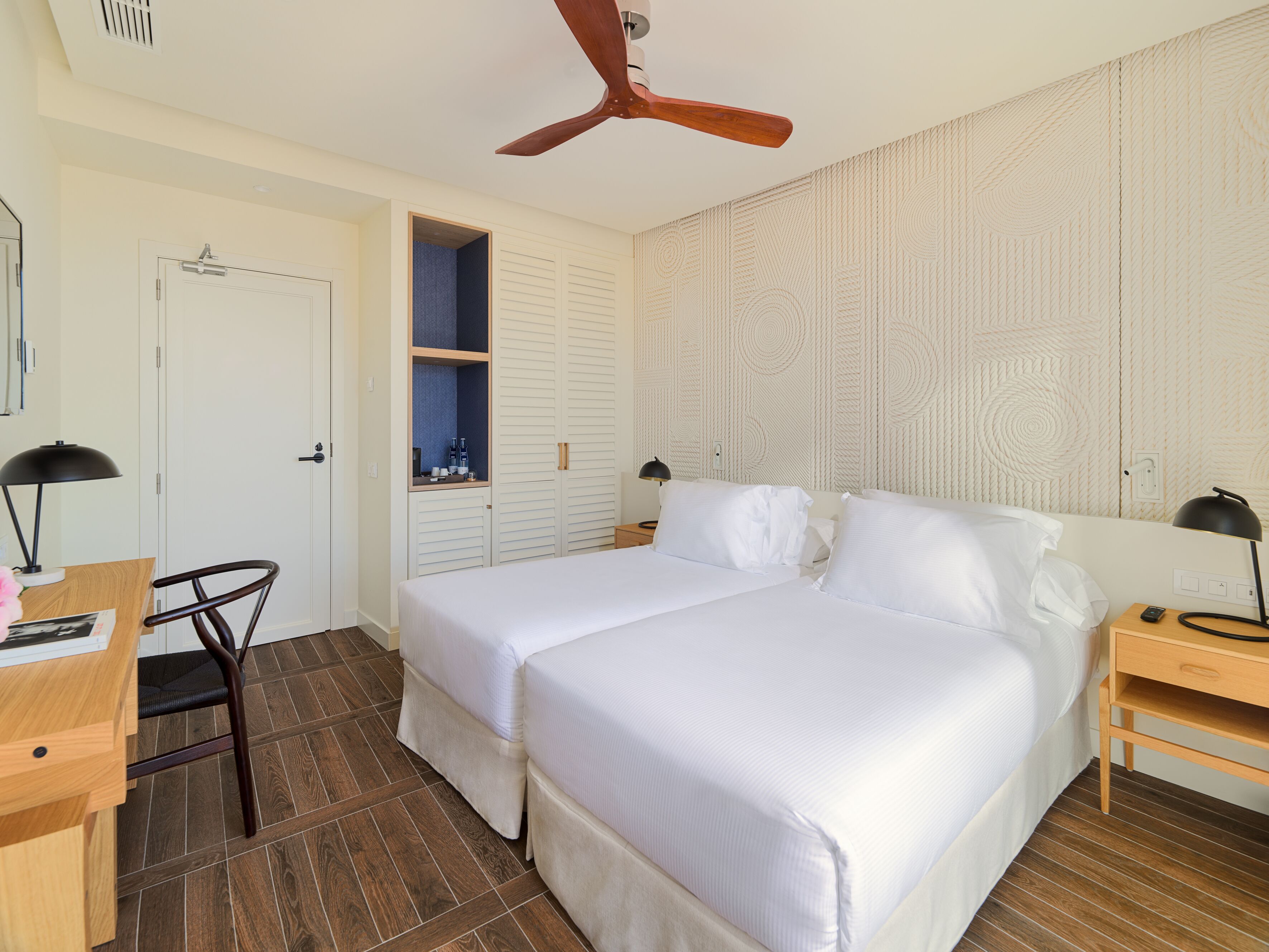 Une chambre d’hôtel avec deux lits, un bureau, une chaise, une armoire et un ventilateur de plafond.