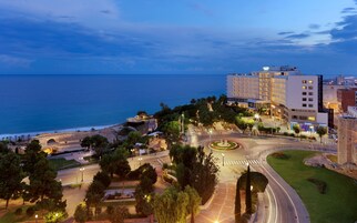 A coastal cityscape with a prominent hotel featuring multiple floors and balconies, a roundabout, and a view of the sea.