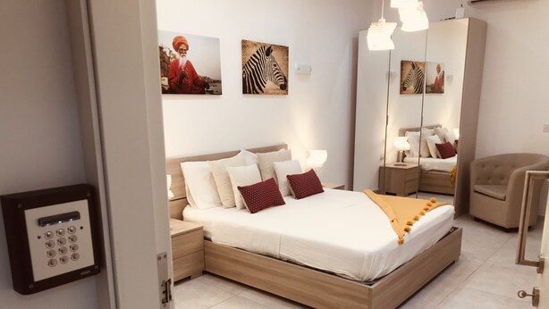Un dormitorio moderno con una cama de madera, ropa de cama blanca y una mesita de noche con lámpara. Se observan obras de arte colgadas en la pared y un espejo.