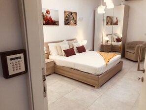 A modern bedroom with a wooden bed, white bedding, and a bedside table with a lamp. There are wall-mounted artworks and a mirror. - LA88 Boutique (St. Paul's Bay)